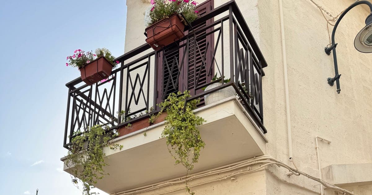 A white house on a southern-Italy street, with a flower-draped wrought-iron balcony against a blue sky.